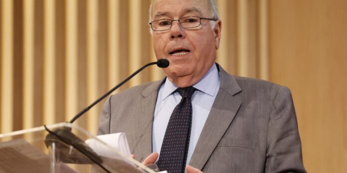 Older man in a gray suit and polka-dot tie speaks into a microphone at a lectern.