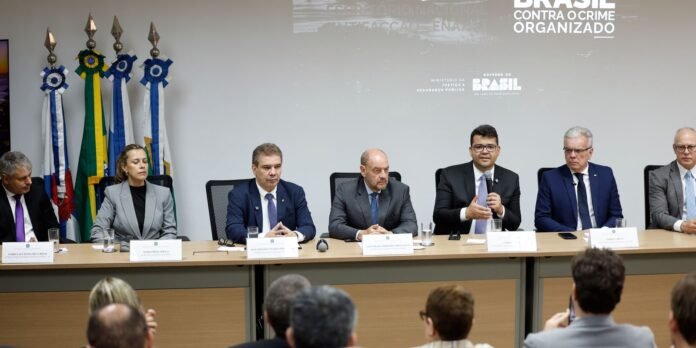 Panel of seven suited officials seated at a long conference table with microphones and nameplates, flags to the left, and a grey wall displaying Brasil contr a crime organizado text in the background.
