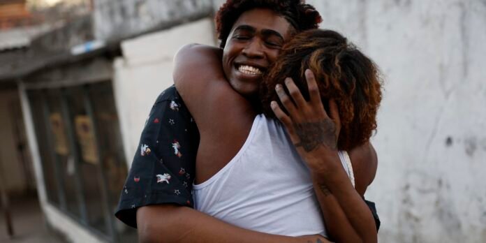 Two friends share a warm hug outdoors, smiling with eyes closed.