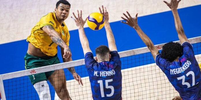 Volleyball players in mid‑game jump at the net, one in yellow and green spikes while two blue‑jerseyed blockers reach up to block the ball at the net.