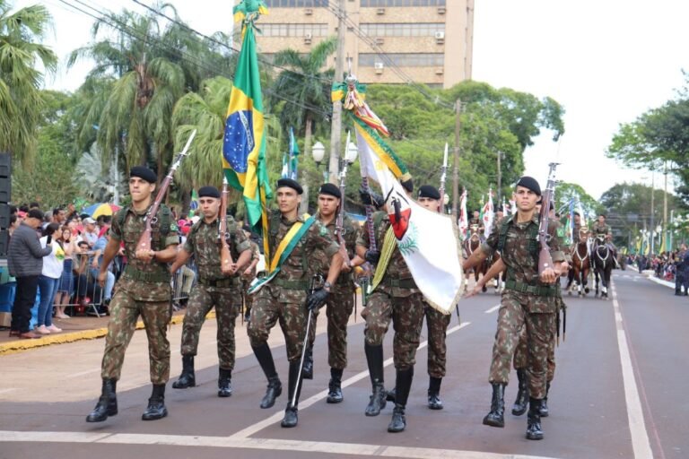 Soldiers in camouflage march down a city street in a parade, carrying rifles and the Brazilian flag, with spectators along the curb.