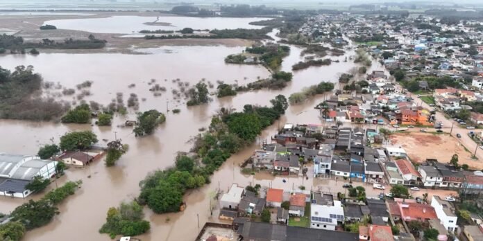 Aerial view of a flood: brown water covers streets and fields, with trees and houses partially submerged near a residential area.