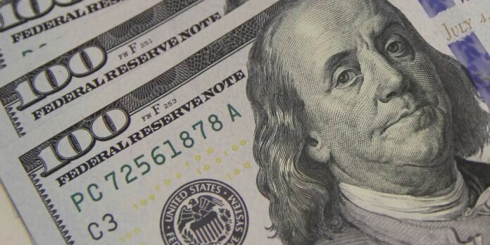 Close-up of several overlapping $100 bills, featuring Benjamin Franklin's portrait and the '100' numerals and seals visible.