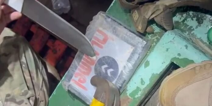 Close-up of a person in camouflage using a knife to pry open a weathered metal box mounted on a green surface.
