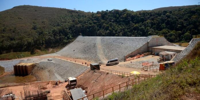 Industrial quarry site with graded gravel slopes, a road, a white van, and temporary workers’ huts amid surrounding hills and trees.