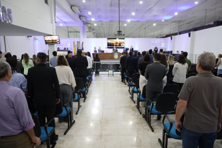 Large conference hall with a panel of speakers at a long desk on a stage, audience standing and facing forward.