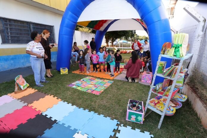 Children form a circle under a large blue inflatable arch at an outdoor kids’ activity area, with colorful mats and books nearby.