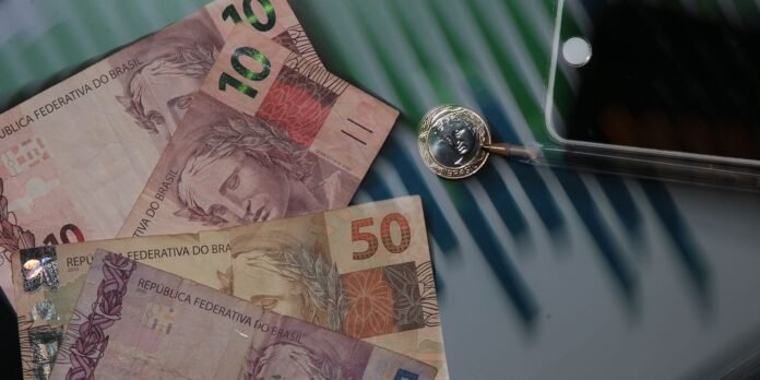 Brazilian real banknotes (R$100 and R$50) spread on a glass table near a silver coin and a smartphone edge, suggesting a payment scene.
