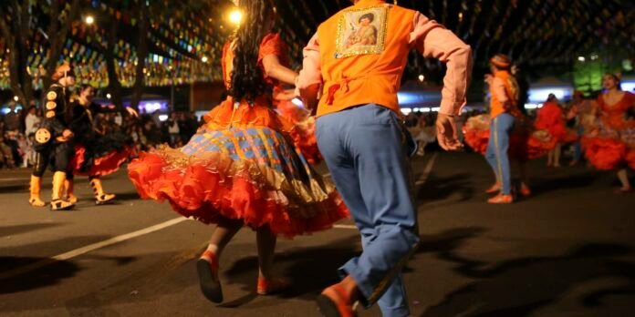 Dancers in bright orange and blue costumes perform in a festive street parade at night before a crowd under colorful lights.