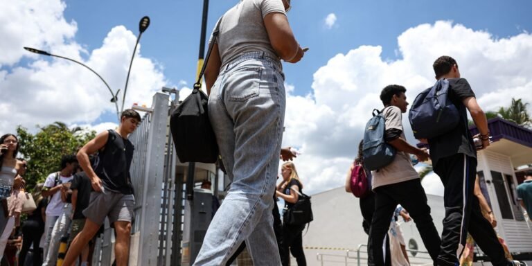 Group of young people walking outdoors on a sunny day, backpacks and casual clothes, near a metal fence gate.