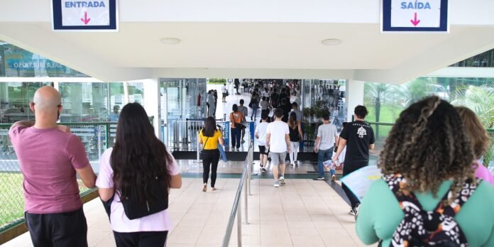 Crowded indoor walkway with people walking toward glass doors under signs reading Entrada and Saída, a transit or hospital-like entrance.