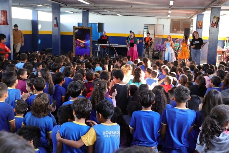 Large group of schoolchildren in blue uniforms watching a band perform on a stage in a gymnasium.