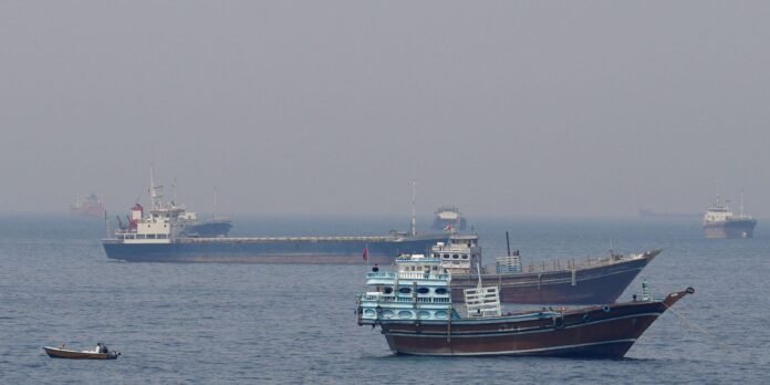 Foreground wooden boat with blue cabins and rigging, on calm sea with several larger vessels in the midground and hazy distant ships in the background.
