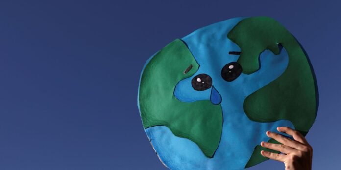Paper cutout of Earth with a sad face and tear, held up by a hand against a clear blue sky.