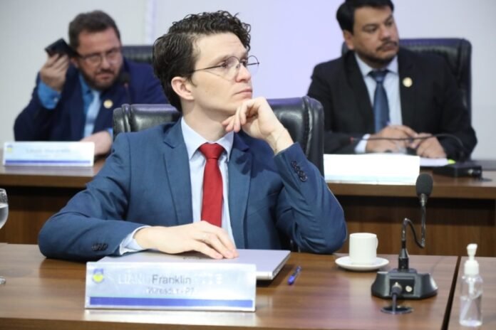 Man in a blue suit and red tie sits at a conference table, hand on chin, listening attentively with two colleagues in the background.