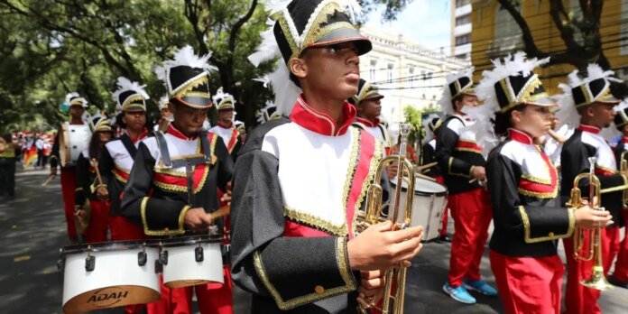 Young marching band members in black, white, and red uniforms with gold trim perform in a parade, trumpet players at the front beside drum lines under trees