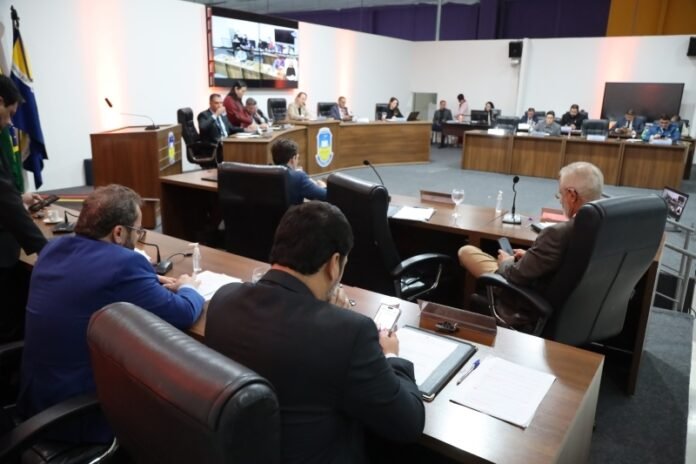 Formal council or board meeting in a large chamber with officials seated around curved wooden desks and microphones on the desks.