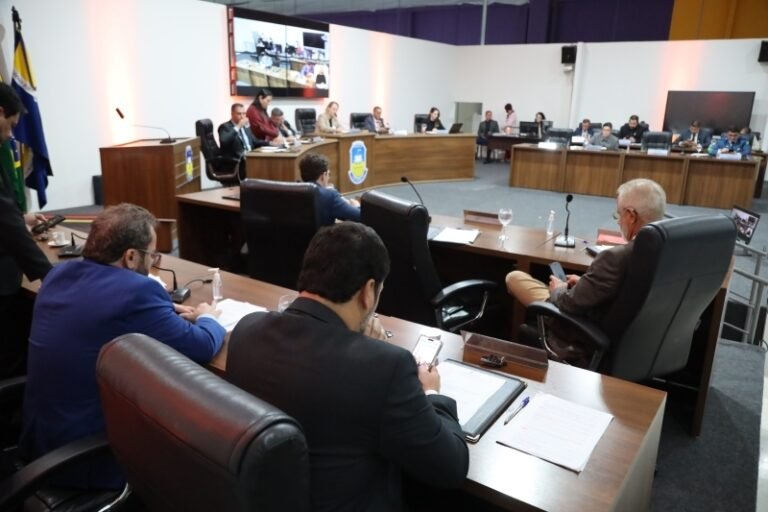 Formal council or board meeting in a large chamber with officials seated around curved wooden desks and microphones on the desks.