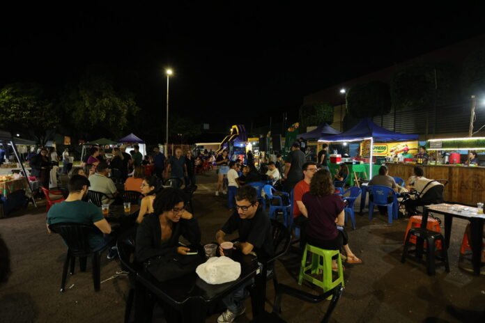 Nighttime outdoor market with many people seated at plastic tables under tents, food stalls in the background.
