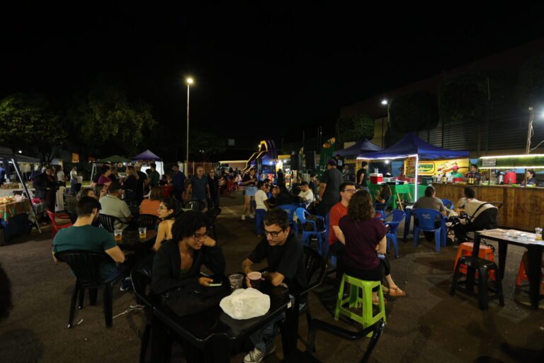 Nighttime outdoor market with many people seated at plastic tables under tents, food stalls in the background.