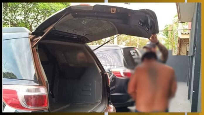 Shirtless man with blurred face stands near an open SUV trunk, police car in the background.