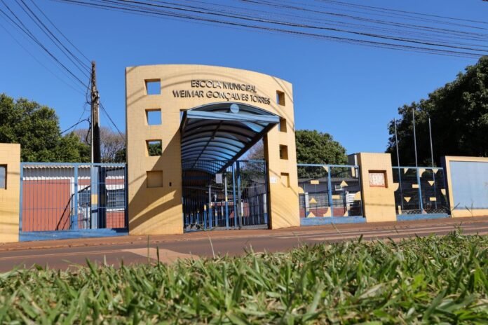 Entrance to Escola Municipal Weimar Gonçalves Torres with a curved glass canopy and blue gates under a clear blue sky.