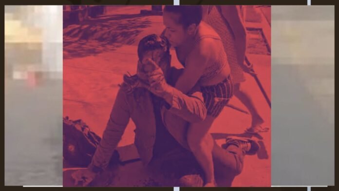 Two men outdoors; one kneels beside the other and cups his face as the second rests on the ground.