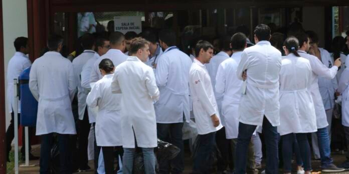 Group of people in white lab coats gathered outside a building, chatting, with a sign reading 'Sala de Espera Inicial' in the background.