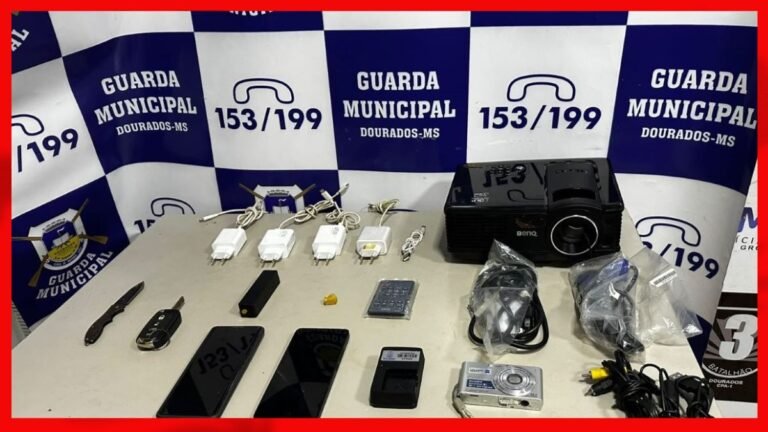 Table of seized items displayed in front of a blue-and-white banner reading Guarda Municipal Dourados-MS, including knives, phones, chargers, a projector, camera, and cables.