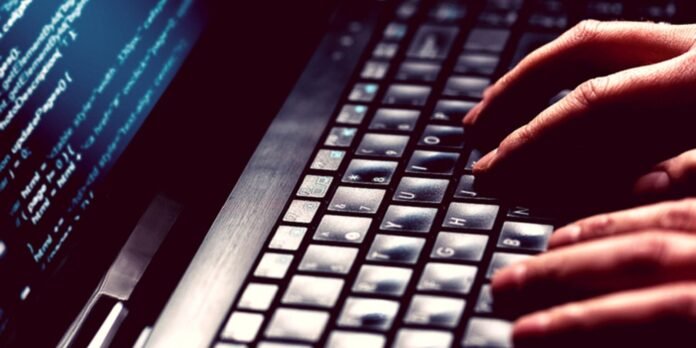 Close-up of hands typing on a keyboard beside a monitor displaying blue code lines.