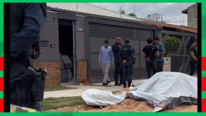 Police officers and investigators stand near a gated house as a body lies covered by a white sheet in the yard.