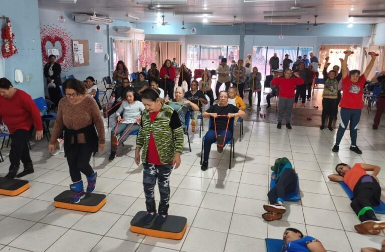 Group of seniors in a community center doing a fitness class with stepping platforms and resistance bands, while others lie on mats in the background.