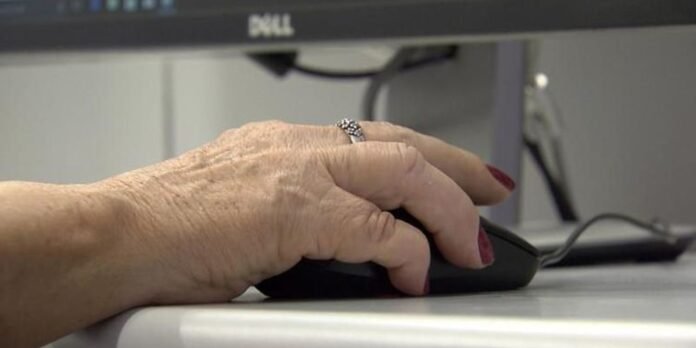 Close-up of an elderly hand with red nail polish on a computer mouse at a desk, with a Dell monitor in the background (informational).