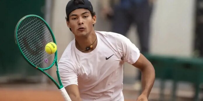Tennis player in a white shirt and backward cap swings a green racket at a yellow ball on court