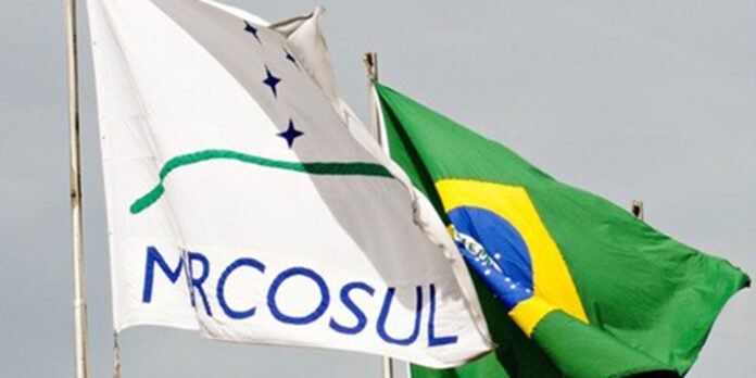 Two flags waving in the wind: a white flag with blue letters and the Brazilian flag on adjacent poles.