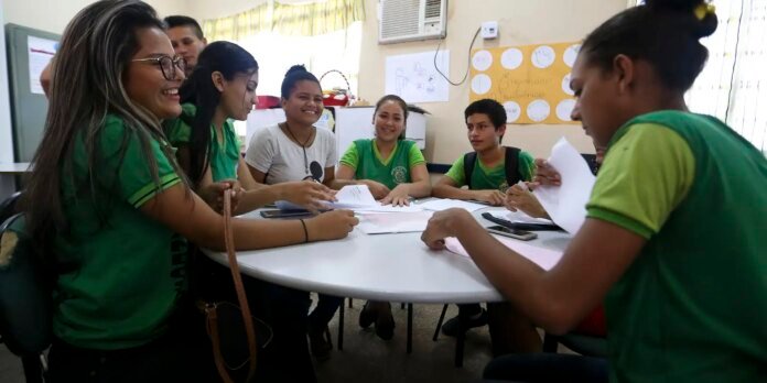 Group of students in green shirts sit around a round table, smiling and working on papers in a classroom.