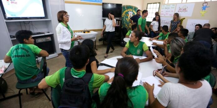 Teacher leads a workshop in a classroom with students in green shirts seated around white tables, watching a presentation on a screen at the front.