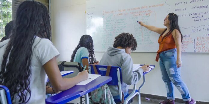 Female teacher in orange top points to a whiteboard full of notes while students sit at blue desks taking notes in a classroom.