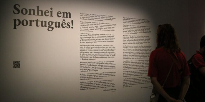 Gallery wall: 'Sonhei em português!' in large type with dense text blocks and a QR code; visitors reading nearby.