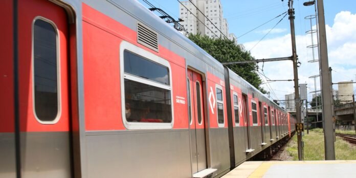 Red urban metro train at an outdoor platform in São Paulo, with overhead power lines and tall buildings in the background.