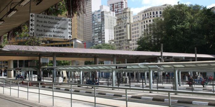 Transit plaza with glass-covered shelters along a curb, tall buildings in the background and people waiting or walking nearby; urban scene.