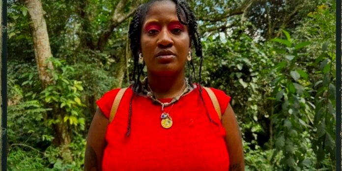 Woman with braided hair in a bright red top, wearing necklaces, standing in a dense green forest.