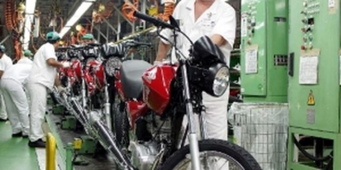 Factory assembly line with workers in white uniforms assembling red motorcycles; a technician adjusts a bike near green machinery in a bright industrial setting.