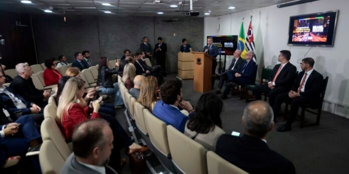 Speaker at a podium addressing an audience in a conference room, with a panel of officials to the right and flags behind.