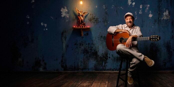 Smiling man in a cap sits on a stool with an acoustic guitar in a worn blue-walled room, a small statue on a lit shelf behind him.
