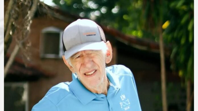 Older man wearing a light blue polo and gray cap, smiling outdoors with greenery in the background.