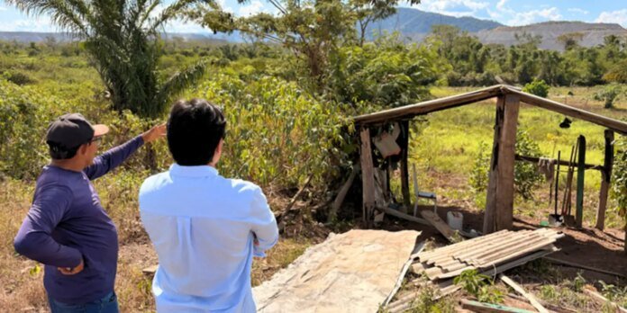 Two men stand in a sunlit rural field, one pointing toward a weathered wooden shelter with corrugated metal sheets and scattered debris. In the background are trees and distant hills or mountains.