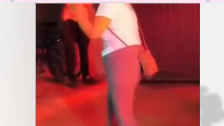Singer performing on a stage with red lighting, holding a microphone in a white shirt and pink pants stance.