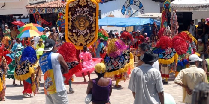 Vibrant street carnival with dancers in colorful feathered capes and fringe, watching crowd in a sunny plaza.