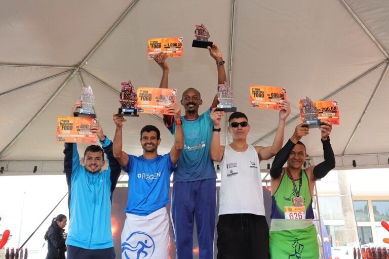 Group of six runners on a stage raising trophies and orange prize certificates under a tenty setup, celebrating wins.